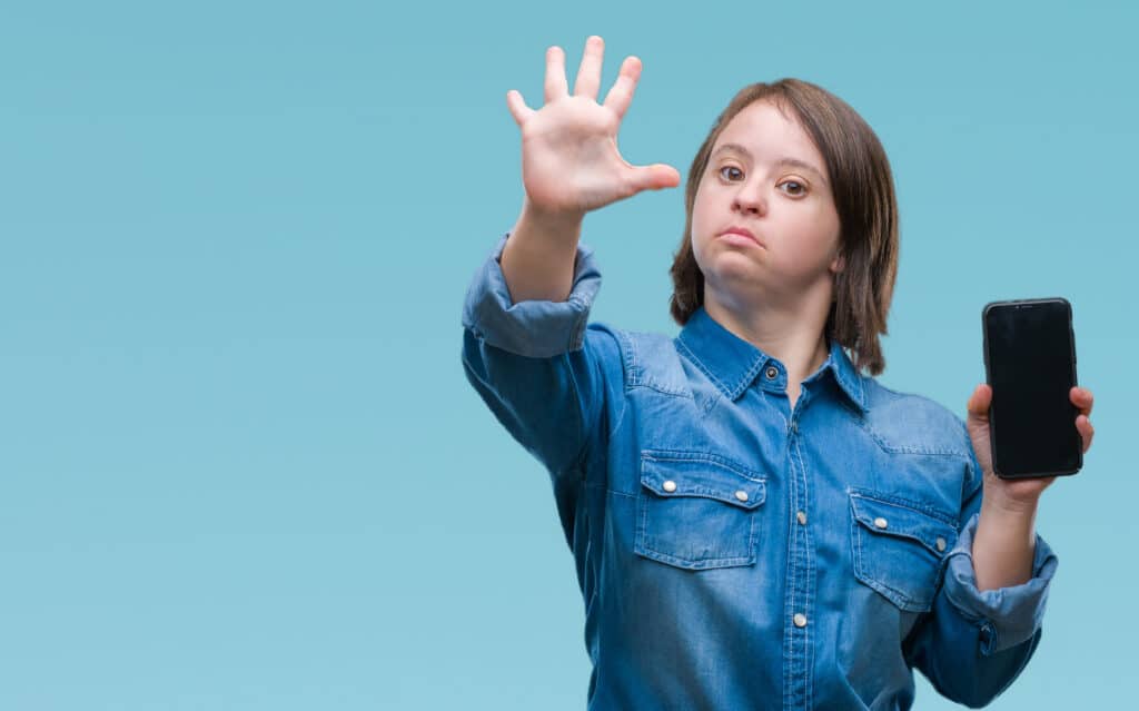 Girl with brown hair and blue jeans shirt raising her hand facing forward. In the other hand she holds a phone.